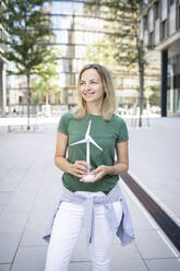 Mid adult woman looking away while holding wind turbine standing on footpath in city - HMEF01114