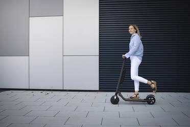 Mid adult woman driving electric push scooter on footpath by metal wall - HMEF01115