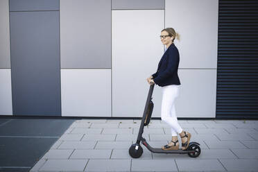 Woman driving electric push scooter on footpath in city - HMEF01116