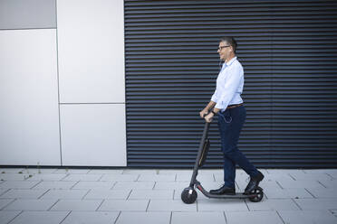 Businessman listening music through earphone standing on electric push scooter - HMEF01121