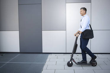 Mature businessman listening music through earphone standing on electric push scooter - HMEF01122