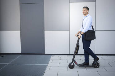 Mature businessman looking away listening music through earphone standing on electric push scooter - HMEF01123