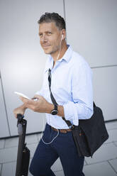 Mature businessman using smart phone listening music through earphone standing on electric push scooter - HMEF01124