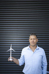 Businessman holding wind turbine while standing against metal wall - HMEF01128