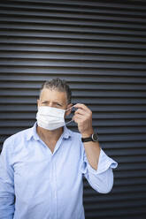 Mature man wearing protective face mask while standing against metal wall - HMEF01129