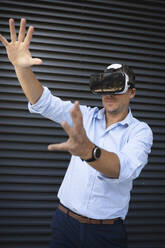 Businessman wearing virtual reality goggles while standing against metal wall - HMEF01132