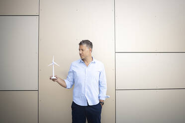 Businessman with hands in pockets holding wind turbine standing against wall - HMEF01134