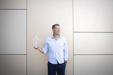 Mature man looking away while holding wind turbine standing against wall - HMEF01136