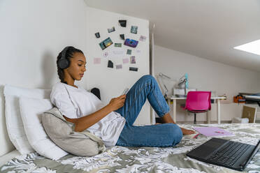 Junges Mädchen hört Musik, während sie ihr Smartphone benutzt und zu Hause im Schlafzimmer sitzt - MGIF01021