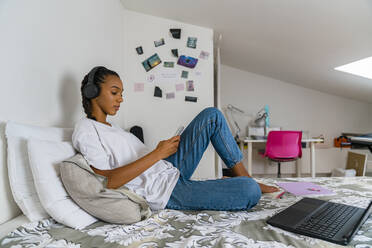 Young girl listening music while using smart phone sitting at home in bedroom - MGIF01021