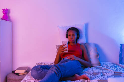 Teenager-Mädchen auf dem Bett liegend, das sein Smartphone benutzt, um zu Hause über Kopfhörer Musik zu hören - MGIF01043