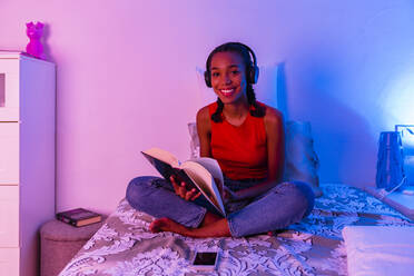 Smiling girl reading book with headphone sitting on bed at home - MGIF01048