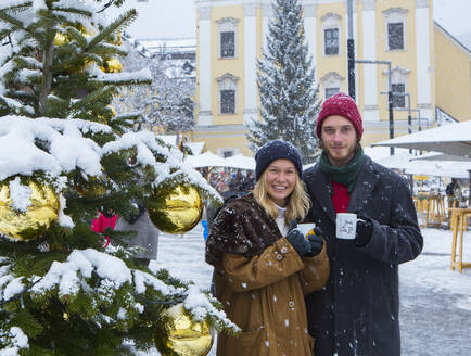 Young couple holding cups with mulled wine while standing by Christmas tree in city - WWF05449