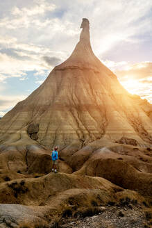 Rückenansicht eines männlichen Touristen in Sommerkleidung, der auf dem ausgetrockneten Boden in Bardenas Reales steht und die herrliche Aussicht auf eine natürliche Felsformation genießt - ADSF16789