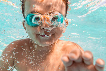 Unterwasseransicht des positiven Jungen mit Schwimmbrille, der in sauberem Wasser im Pool schwimmt, während er am Wochenende im Sommer Spaß hat - ADSF16818