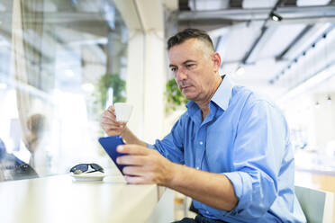 Männlicher Unternehmer, der ein Mobiltelefon benutzt, während er in einem Café Kaffee trinkt - WPEF03433