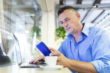 Handsome male professional using mobile phone and laptop while sitting at cafe - WPEF03435