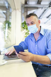 Männlicher Unternehmer trägt einen Gesichtsschutz, während er in einem Café sein Smartphone benutzt - WPEF03451
