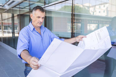 Confident male entrepreneur looking at floor plan while standing in city - WPEF03457