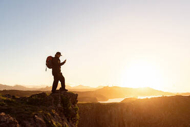 Seitenansicht eines männlichen Forschers mit Rucksack, der auf einem felsigen Hügel steht und ein Foto von einer spektakulären Berglandschaft bei Sonnenuntergang macht - ADSF16819