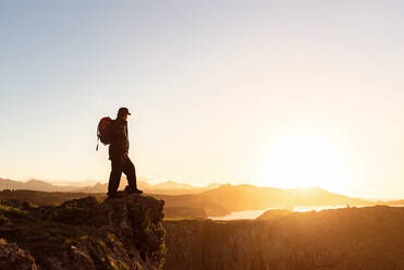 Seitenansicht eines anonymen männlichen Wanderers mit Rucksack, der auf einem Felsen in den Bergen steht und den herrlichen Sonnenuntergang im Urlaub genießt - ADSF16820