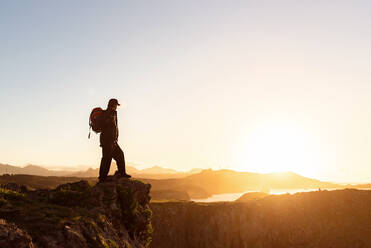 Side view of anonymous male hiker with backpack standing on rock in mountains and enjoying amazing sundown during vacation - ADSF16820