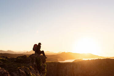 Side view of anonymous male hiker with backpack sitting on rock in mountains and enjoying amazing sundown during vacation - ADSF16821