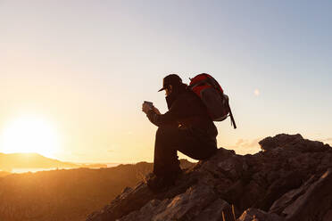 Side view of unrecognizable male hiker sitting on rock in highlands and taking photo while admiring sunset - ADSF16822