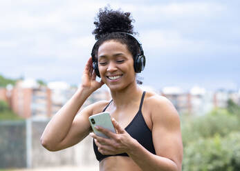 Delighted black female athlete in sports bra standing in city and enjoying songs with closed eyes in wireless headphones during training - ADSF16877
