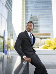 Side view of confident black male executive manager in formal suit sitting on fence in megalopolis and looking away - ADSF16885