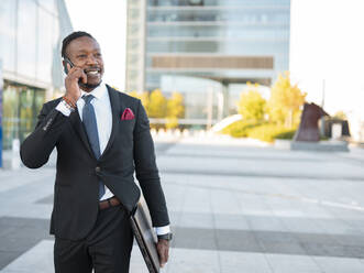 Zufriedener afroamerikanischer männlicher Unternehmer in formellem Anzug, der mit einem Laptop die Straße entlangläuft und sich über ein Smartphone unterhält, während er ein Geschäftsprojekt bespricht und wegschaut - ADSF16886