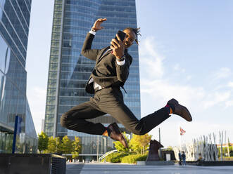 Side view of carefree black male entrepreneur in classy suit and with smartphone jumping on street in downtown and looking at camera - ADSF16887