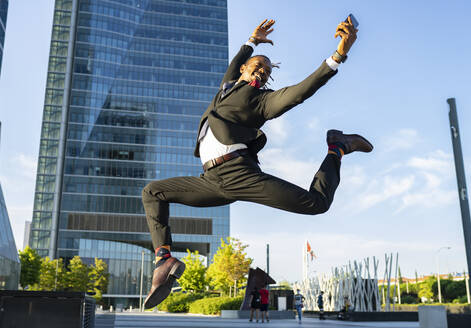 Seitenansicht eines unbekümmerten schwarzen Unternehmers im eleganten Anzug und mit Smartphone, der in der Innenstadt auf die Straße springt und wegschaut - ADSF16888