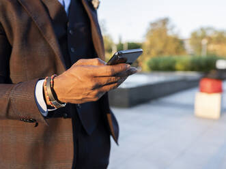 Unrecognizable ethnic businessman in stylish suit standing in city and surfing Internet on smartphone while working remotely on project - ADSF16893