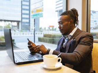 Side view of concentrated African American businessman in suit sitting at table with coffee in cafe and browsing smartphone while working on remote project - ADSF16895