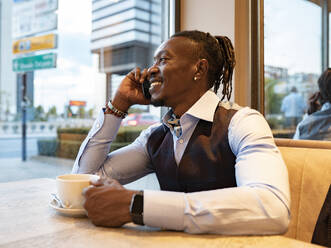 Positiver afroamerikanischer männlicher Unternehmer in stilvollem Outfit, der mit einer Tasse Kaffee in einem Café am Tisch sitzt und mit dem Handy spricht, während er aus dem Fenster schaut - ADSF16896