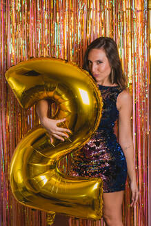 Side view of young female in bright festive outfit embracing golden balloon in shape of number 2 against colorful tinsel while celebrating New Year holiday - ADSF16901