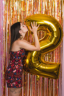 Side view of young female in bright festive outfit kissing golden balloon in shape of number 2 against colorful tinsel while celebrating New Year holiday - ADSF16906