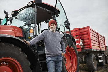 Lächelnder reifer Landwirt steht mit der Hand in der Tasche vor einem roten Traktor auf einem Bauernhof - ZEDF03958