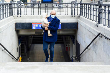 Mann mit Gesichtsmaske trägt Baby beim Gehen auf einer Treppe in der Stadt - PGF00147