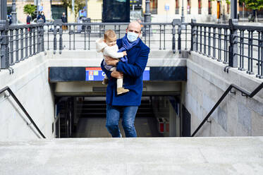 Vater mit Gesichtsmaske trägt seinen Sohn beim Gehen auf einer Treppe in der Stadt - PGF00148