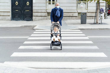 Mann mit Gesichtsmaske geht mit Kinderwagen über die Straße in der Stadt - PGF00152