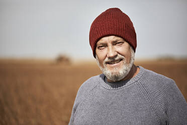 Farmer wearing knit hat smiling while standing at soybean farm - ZEDF03964