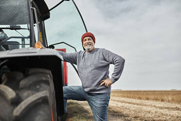 Selbstbewusster Landwirt, der mit der Hand in der Hüfte neben einem Traktor auf einem Bauernhof steht - ZEDF03974