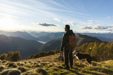 Wanderer mit Hund und Blick auf eine Bergkette bei Sonnenuntergang - MCVF00631