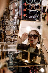 Reflektion einer selbstbewussten Frau, die eine Sonnenbrille im Spiegel eines Modegeschäfts ausprobiert - MASF19966