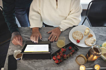Blick von oben auf ein älteres Paar, das ein digitales Tablet benutzt, während es am Esstisch im Wohnzimmer sitzt - MASF19975