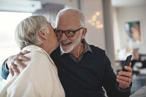 Senior woman kissing partner with smart phone while sitting in living room stock photo