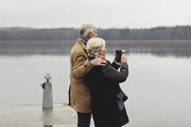 Senior couple photographing while standing by lake against clear sky - MASF19984