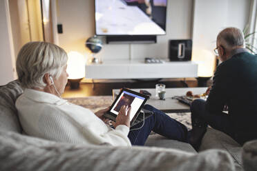 Ältere Frau benutzt ein digitales Tablet, während ihr männlicher Partner auf dem Sofa im Wohnzimmer sitzt - MASF19999